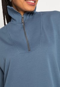 Pullover azul com fecho de meia-zíper em tecido texturizado, gola alta e um zíper prateado estreito. Apresenta uma pala moldada e um acabamento suave.