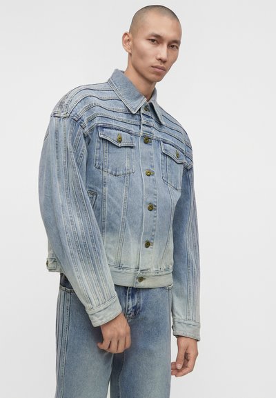 Άνδρας που φοράει φαρδιά ανοιχτό μπλε τζιν μπουφάν με κατακόρυφες ραφές και ταιριαστό τζιν παντελόνι σε μονόχρωμο λευκό φόντο.