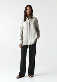 Chemise ample gris clair à boutons, à manches longues, avec une poche poitrine, associée à un pantalon noir large et des sandales plates.