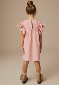 Rosa Baumwollkleid mit Rüschensäumen, Schlitzrücken und floralem Stickereien. Es werden tanfarbene Knöchelstiefel mit Fransenakzenten getragen. Glatte Textur.