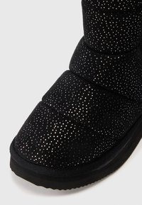 Bota acolchada negra con un pequeño patrón de puntos reflectantes y suela gruesa sobre un fondo claro y liso.
