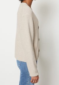 Beżowy sweter robiony na drutach z dekoltem w serek, długimi rękawami, z zapięciem na guziki, prążkowanym dołem i widoczną teksturą. Nosi się go na jasnoniebieskich dżinsach.