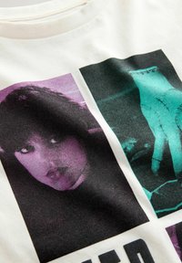 Tricou din bumbac alb-crem, cu două imprimeuri grafice: unul în purpuriu cu fața unei femei și altul în teal cu o mână cusută, având accente negre.