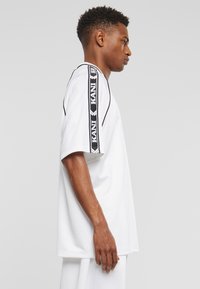 Balta per didelė t-shirt su kontrastingomis juodomis vertikaliomis šoninėmis juostelėmis, ant kurių yra tekstas. Trumpomis rankovėmis, apvaliu kaklu ir lygiu paviršiumi.