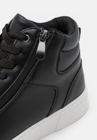 Zapatilla alta de cuero negro con cremallera lateral, parte superior texturizada y suela de goma blanca con acabado suave.