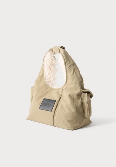 Sac à main en tissu doux beige avec une étiquette noire à l'avant et une doublure blanche duveteuse à l'intérieur de la sangle d'épaule.