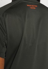 Ciemnozielona koszulka sportowa z teksturowanej tkaniny, z małym pomarańczowym logo "Manchester United" na tylnej części kołnierza.