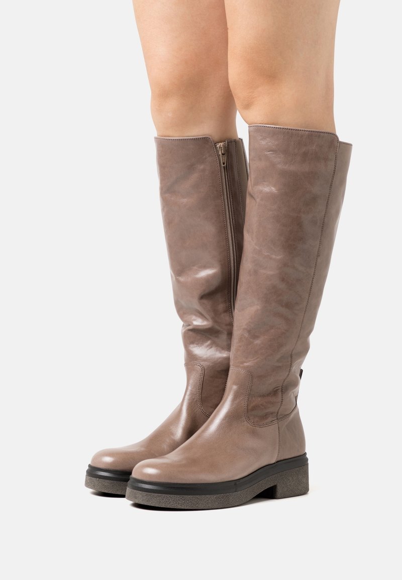 Bottes hautes en cuir taupe avec fermeture éclair latérale et semelle épaisse noire portées par une personne, visibles de mi-mollet vers le bas sur un fond blanc.