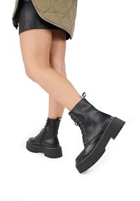 Stivali alla caviglia in pelle nera con lacci, dotati di una suola a piattaforma chunky e dettagli di cucitura bianca. Presentano una texture liscia e un design con punta tonda.