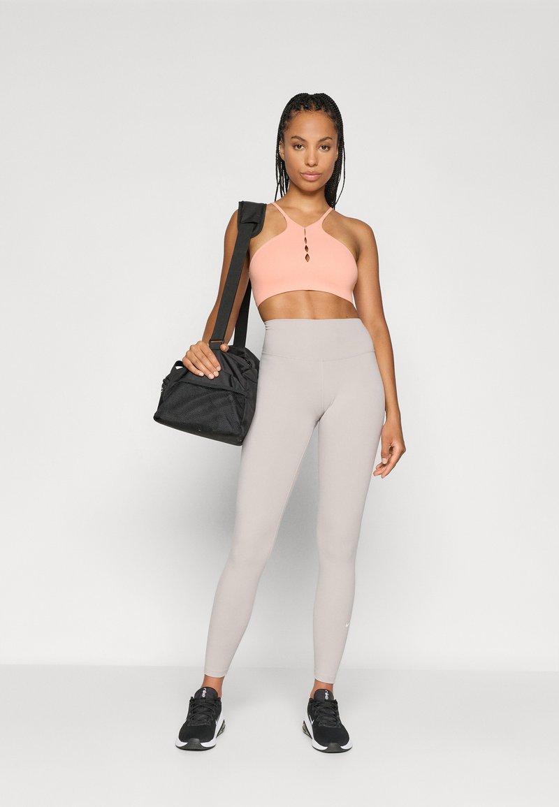 Schwarze Sporttasche, hellrosa Sport-BH mit einem Schlüsselelement und hellgraue, hoch taillierte Leggings. Schwarze Sneakers runden das Outfit ab.