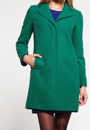 Vrouw draagt een knie-lange groene jas met kraag, met een hand in de zak, over een donkerblauwe jurk en zwarte panty's.