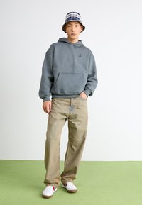 Grå hætte sweatshirt med kængurulomme og sort logo, lysebrune baggy jeans og hvide sneakers med røde og blå accenter.