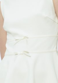 Close-up van een witte mouwloze jurk met twee dunne taillebanden die aan de achterkant zijn gestrikt in kleine strikjes.