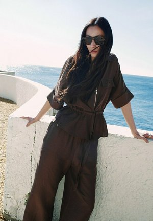 Kobieta ubrana w luźny, ciemnobrązowy strój i okulary przeciwsłoneczne opiera się o biały betonowy parapet nad oceanem w słoneczny dzień.