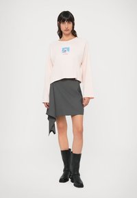 Lichtroze cropped top met lange mouwen en een grafisch ontwerp in het midden, gecombineerd met een grijze asymmetrische rok en zwarte kniehoge laarzen.