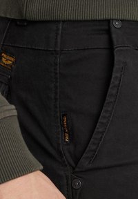 Črne denim hlače z gladko strukturo, z oranžno oznako in gumbnim zapiranjem. Ob strani je vidna zelena rebrasta tkanina.