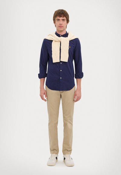 Camisa azul marinho de botões combinada com calças cáqui bege. Suéter grosso de malha creme pousado sobre os ombros. Ténis brancos com detalhes pretos.