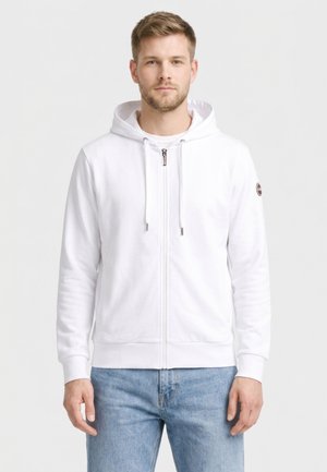 Jonge man die een witte hoodie met ritssluiting en trekkoorden draagt, en lichtblauwe jeans, staand tegen een effen lichtgrijze achtergrond.