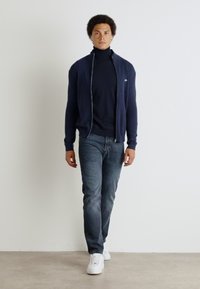 Marinblå zip-up kofta över en svart polo, parats med grå jeans och vita sneakers. Slät tyg, skräddarsydd passform och minimalistisk design.