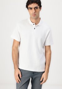 Hvid polo skjorte med struktureret stof, korte ærmer og en tre-knaps lukning med sorte knapper. Bæres med mørke jeans.