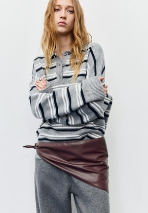 Junge Frau mit langen blonden Haaren, die einen grau-navy gestreiften Pullover und graue Hosen mit asymmetrischem bordeauxrotem Lederrock-Detail trägt.