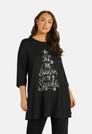 Mujer que lleva una túnica negra de manga larga con texto plateado de lentejuelas formando la forma de un árbol de Navidad que dice "'Tis the season to sparkle."