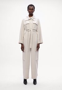 Bež jumpsuit s velikim ovratnikom, dvije prsne džepove, odvojivim pojasom, širokim nogavicama i bočnim džepovima. Model nosi crne gležnjače.