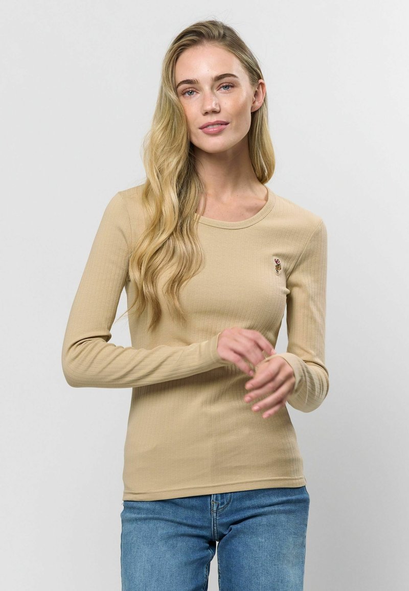 Beige, hosszú ujjú bordás kötött póló, kerek nyakkivágással és egy kis hímzett mintával a mellrészen. Kék farmernadrággal párosítva.