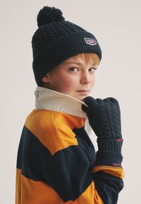 Marineblauw gebreide muts met een pompon, bijpassende handschoenen en een gestreept geel-blauw shirt met kraag, met textuurpatronen en merkdetails.