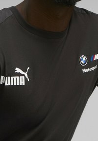 Černé bavlněné tričko s krátkým rukávem. Obsahuje bílé logo Puma a BMW Motorsport. Hladká textura s kulatým výstřihem.