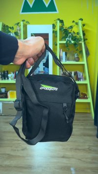 Svart tygaxelväska med justerbara remmar, framficka med dragkedja och grön logotyp. Kompakt design som passar för dagligt bruk.