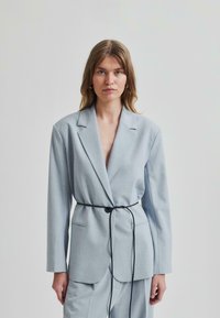 Svijetloplava sako jakna s dubokim V-izrezom, dugim rukavima i detaljem crnog remena u struku. Izrađena od glatkog materijala s minimalnom teksturom.