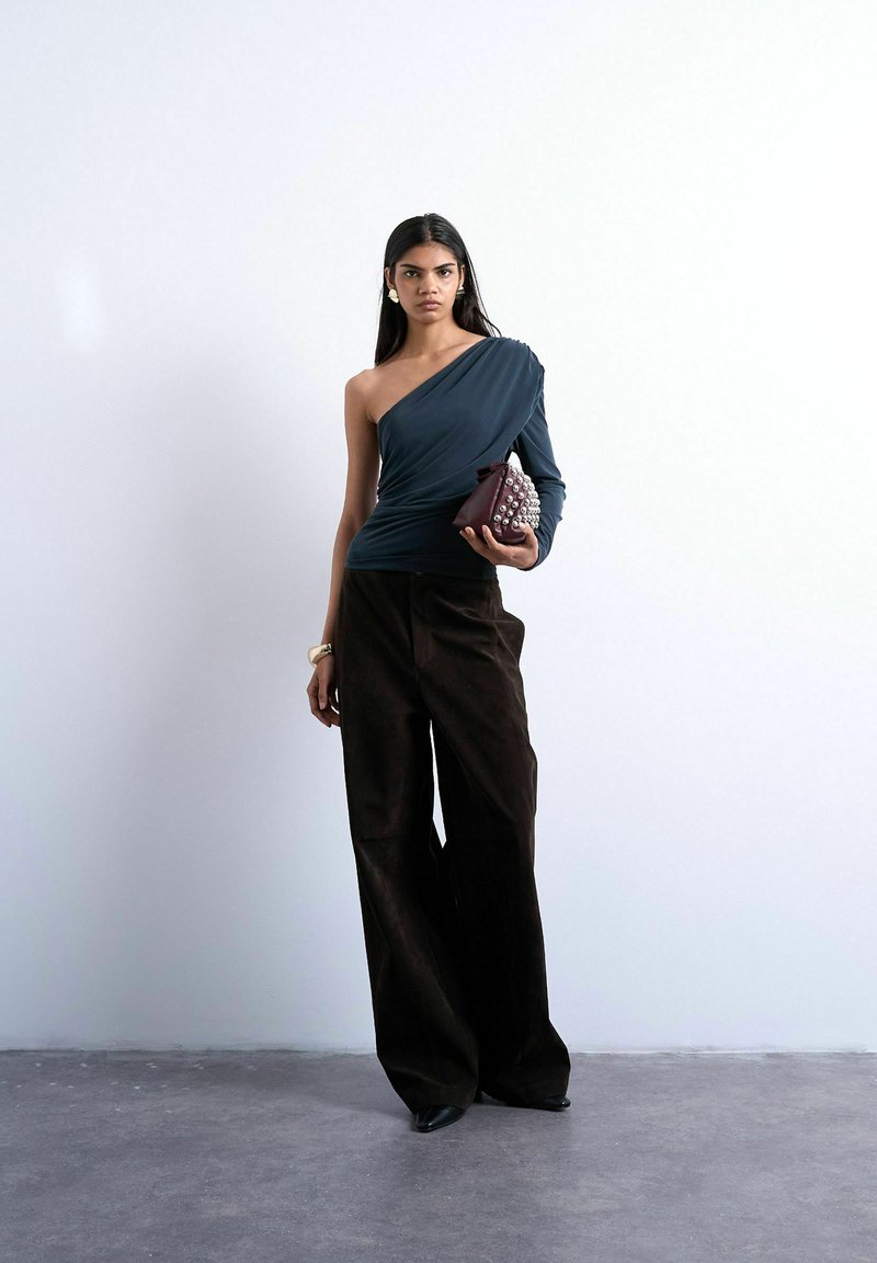 Femme portant un haut noir à une manche, un pantalon noir à jambes larges et des chaussures pointues, tenant une pochette bordeaux cloutée contre un mur uni.