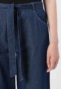 Calças de ganga de um azul escuro com um cinto de tecido atado à cintura e um bolso lateral visível, usadas por uma pessoa com a mão direita descontraída.