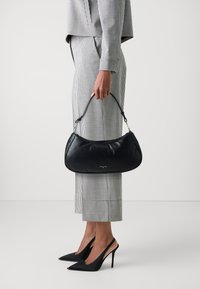 Borsa a tracolla in pelle nera con una texture liscia, forma curva e manico corto, tenuta da una persona in un completo a quadri e tacchi alti.