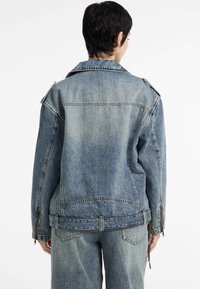 Persoon gezien van achteren, met een blauwe denimjas en bijpassende jeans, tegen een effen witte achtergrond.