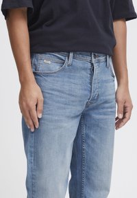 Light blue denim jeans with a straight cut, featuring a small brand label on the front pocket, and a subtle faded texture.