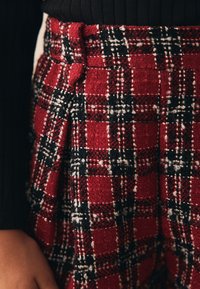 Rote und schwarze kariere Hose aus strukturiertem Stoff, mit hoher Taille, Gürtelschlaufen und Falten für zusätzliche Details.