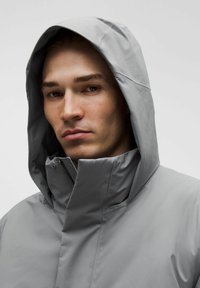 Noor mees kannab heledat halli kapuutsiga jakki, millel on tõmblukk ja kõrge kaelus, vaatab otse ette neutraalse näoilmega, lihtne taust.