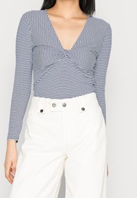 Långärmad randig topp med djup V-ringning, twistdetalj fram, tillverkad av mjukt tyg, kombinerad med högmidjade vita byxor.