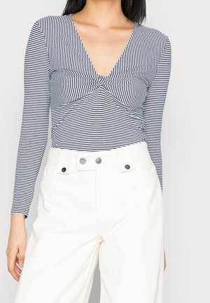Langärmliges gestreiftes Oberteil mit tiefem V-Ausschnitt und Twist-Detail an der Vorderseite, aus weichem Stoff, kombiniert mit hochgeschnittenen weißen Hosen.