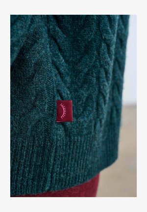 Dunkelgrüner Strickpullover mit strukturierter Zopfmuster-Optik. Verfügt über ein kastanienbraunes Etikett mit rosafarbenem grafischem Akzent in der Nähe des Saums.