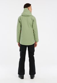 Giacca impermeabile verde con cappuccio, polsini regolabili e tasche, abbinata a pantaloni neri. Texture liscia e vestibilità rilassata.