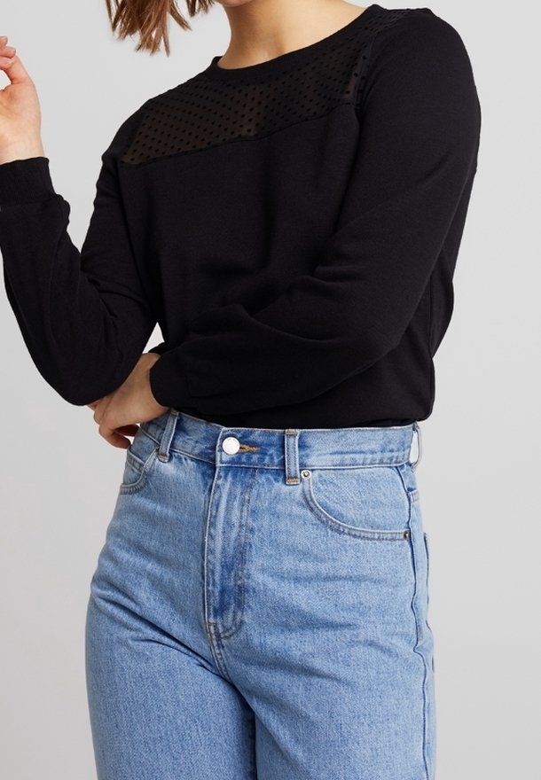 Femme portant un haut noir à manches longues avec un empiècement en tulle à pois transparent et un jean taille haute bleu clair sur un fond uni.