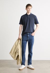 Cămașă polo bleumarin, blugi, adidași albi cu tălpi maronii deschise și o jachetă bej aruncată peste braț. Ținută simplă și casual.