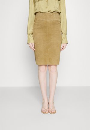 Mujer con una falda lápiz de ante beige, blusa de manga larga de color amarillo claro y sandalias beige, de pie frente a un fondo liso.