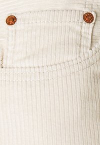 Tela de pana color crema con textura acanalada, que presenta un pequeño bolsillo frontal y detalles de herrajes metálicos.