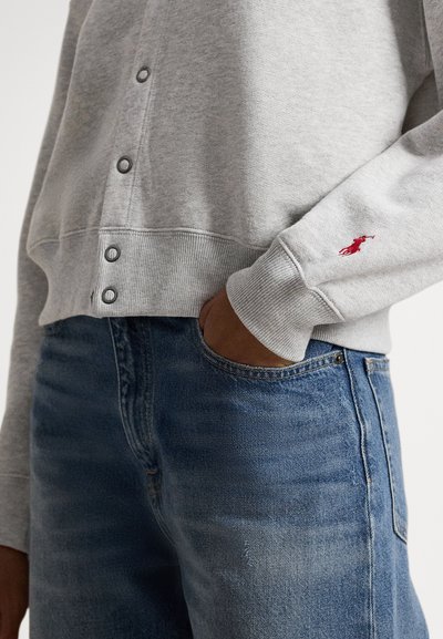 Pessoa vestindo suéter cinza claro abotoado com logótipo vermelho bordado na manga e jeans azuis, mão no bolso.