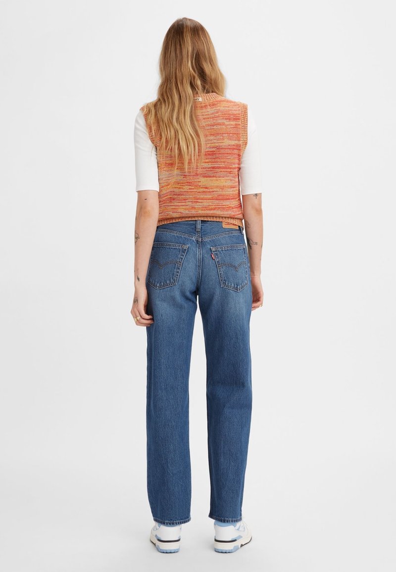 Vue arrière d'une personne portant un gilet en tricot orange raccourci et un jean droit bleu. Des baskets blanches complètent la tenue.