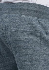 Nahaufnahme von einer blaugrauen Sweatpants mit einer Gesäßtasche und einem gerippten Bund, in dem ein weißes Hemd oberhalb der Taille eingeklemmt ist.